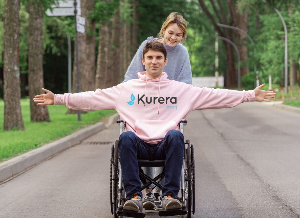 Man som sitter i rullstol och blir knuffad av en kvinnlig assistent. Mannen har en rosa tröja med Kurera Omsorg logotyp.